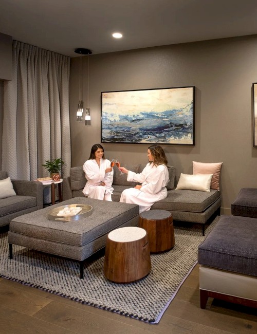 Two people in white robes are sitting and chatting in a cozy lounge with modern furniture, soft lighting, and wall art above, creating a relaxing ambiance.