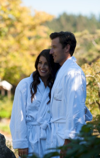 A couple in white robes stands outside in a lush, green garden with trees in the background, smiling and enjoying the scenery.