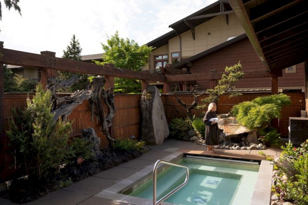 The image shows a small, tranquil outdoor space with a hot tub, surrounded by lush greenery and wooden structures.