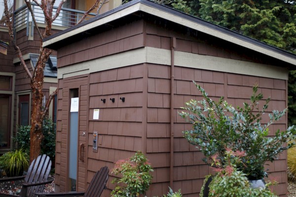 A small wooden building with two brown chairs outside, surrounded by potted plants and trees in a garden setting, next to a larger structure.