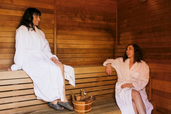 Two women in white robes are relaxing and talking inside a wooden sauna room.