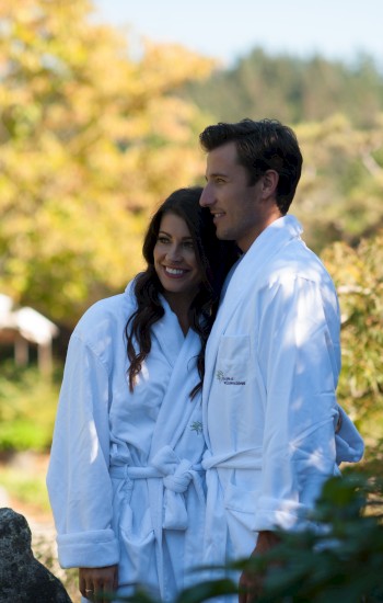 A couple in white robes stands closely together outdoors, surrounded by lush greenery and colorful foliage on a sunny day.