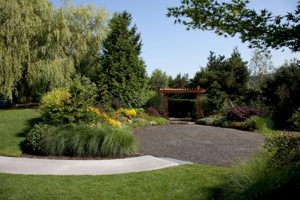 A beautifully landscaped garden with a pathway, flowering plants, tall trees, and a wooden pergola in the background, surrounded by greenery.