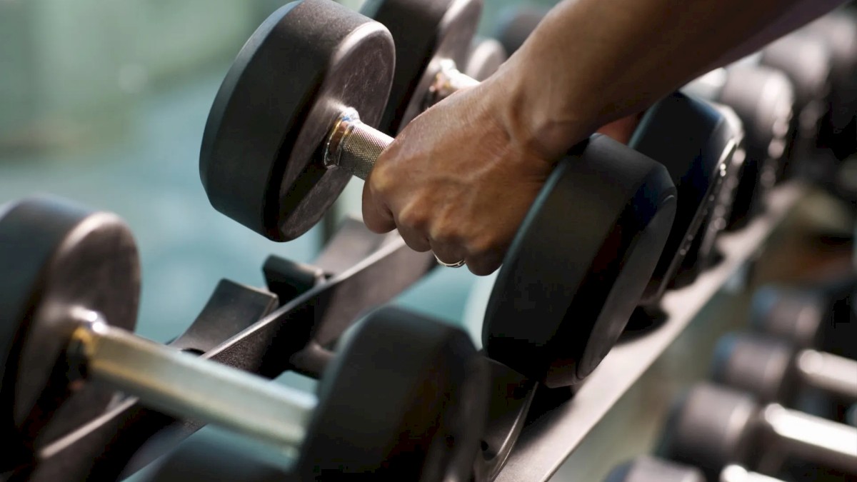 A person is picking up a dumbbell from a rack in a gym, with multiple other dumbbells visible in the image.