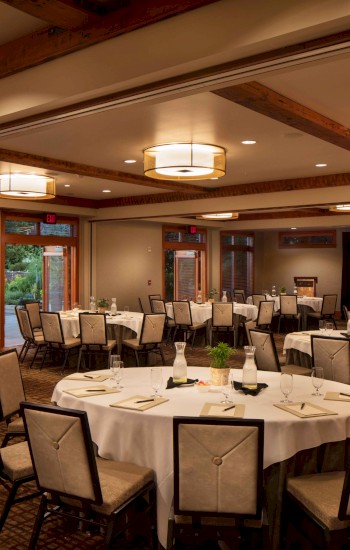 A well-lit conference room with round tables set for a meeting or event, featuring a projector screen, chairs, and a garden view through large windows.