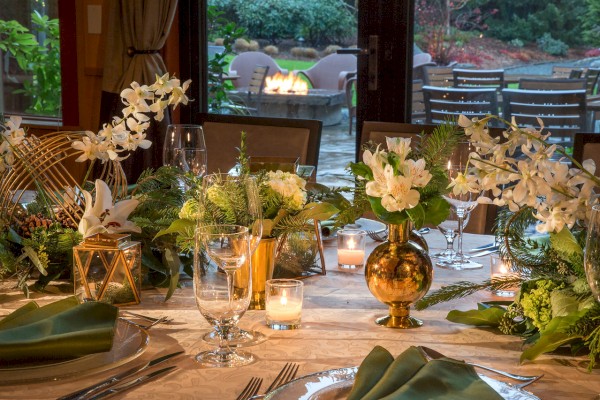 A beautifully set dinner table with green napkins, floral arrangements, and candles, with an outdoor fire pit visible in the background through a window.