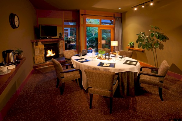 A cozy, well-lit meeting room with a table and five chairs, a fireplace, plants, and a window with a garden view.