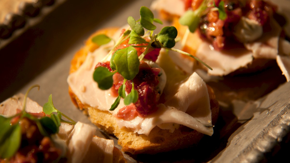 The image shows a tray of gourmet appetizers made of toasted bread topped with sliced meat, a dollop of sauce, and garnished with microgreens.