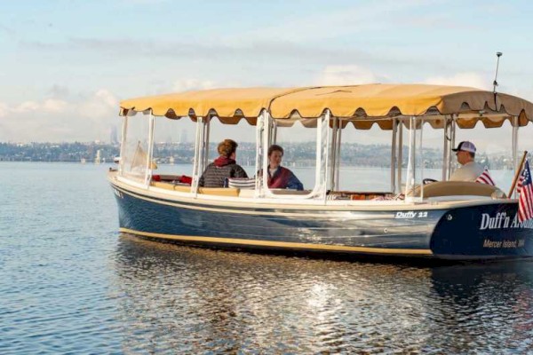 A small boat with canopy and three passengers floating on calm water, with an American flag at the stern and a distant shoreline in the background.