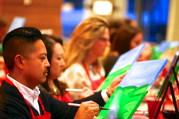 People are seated, painting on canvases with brushes in hand, focused on their work, and wearing aprons in a group setting.