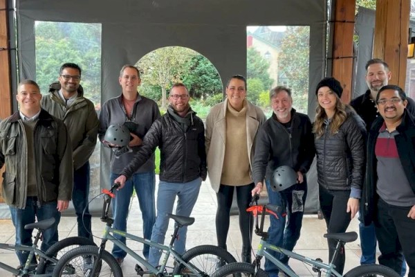 A group of eight people stands in a covered outdoor area, posing with bicycles and helmets. They appear to be enjoying a group activity.