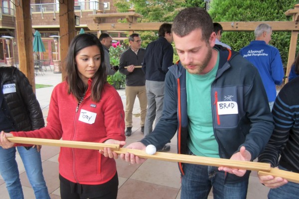 People participating in a team-building activity, using wooden rods to balance a ball. They’re outdoors, wearing casual clothes, name tags visible.