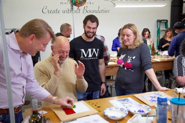 People are gathered around a table engaged in an art activity, with others in the background socializing and creating.