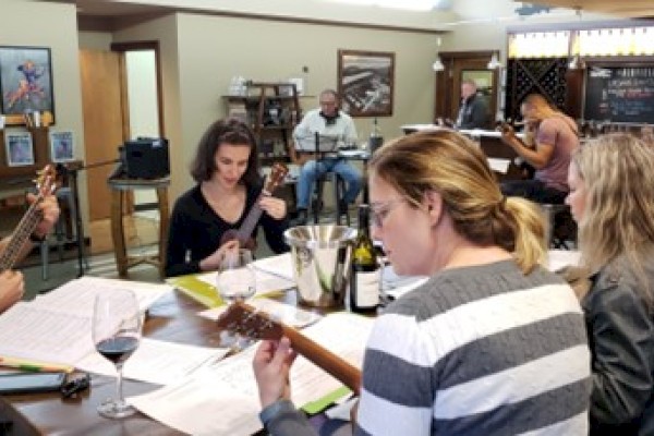 A group of people sits around a table with musical instruments and papers, suggesting a casual gathering or music practice session, always ending the sentence.