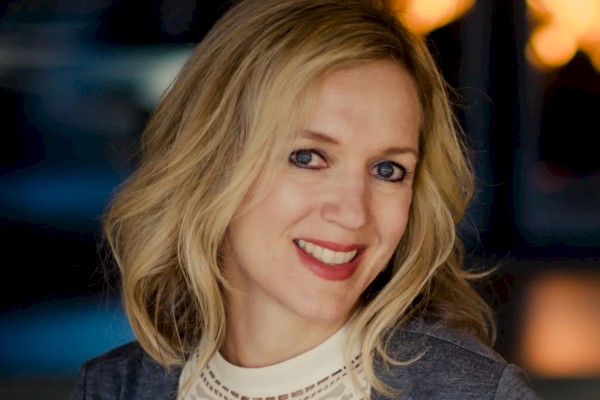 A woman with blonde hair is smiling, wearing a white lace top and a gray blazer, with a blurred, warmly lit background.