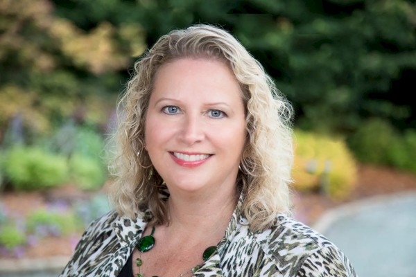 A woman with curly blonde hair is smiling, wearing a patterned blouse and green necklace, with a blurred, green, outdoor background.