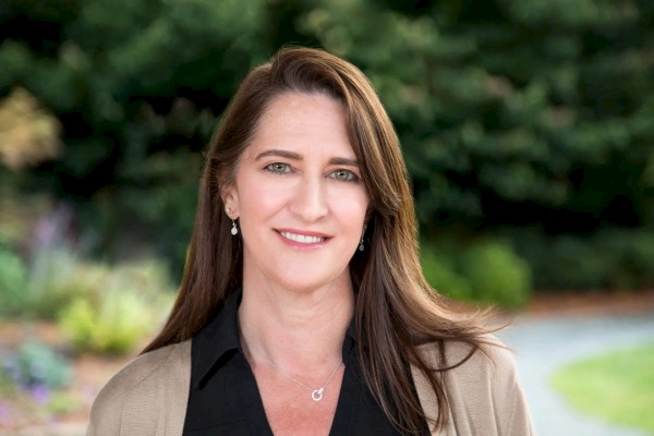 A woman with long brown hair stands outside, wearing a black top and light-colored jacket, smiling in front of greenery.