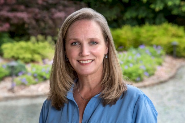 The image shows a woman with long, light brown hair wearing a blue shirt, smiling in an outdoor setting with green plants and a pathway in the background.