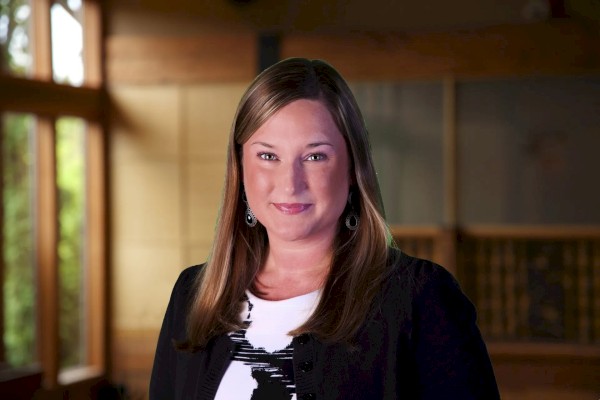 A woman with long hair smiles in an indoor setting, with a softly lit background featuring wooden elements and large windows.