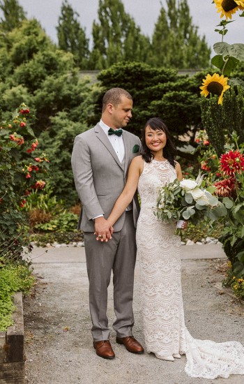 A couple is posing in a lush garden with vibrant flowers. The man is in a gray suit and the woman is in a white wedding dress holding a bouquet.