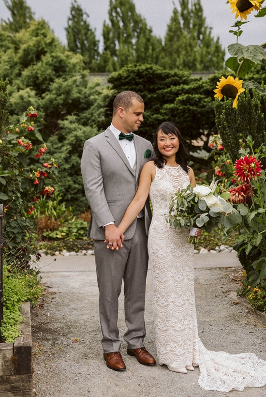 A couple is posing in a lush garden with vibrant flowers. The man is in a gray suit and the woman is in a white wedding dress holding a bouquet.