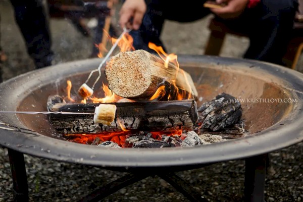 A campfire with people roasting marshmallows over the flames, with logs burning in a metal fire pit, and hands holding roasting sticks.