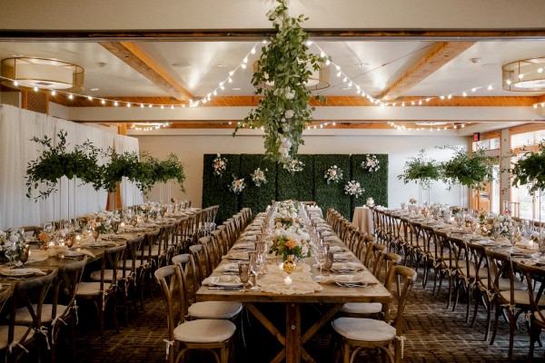 This image displays a beautifully decorated wedding reception venue with long tables, elegant place settings, hanging greenery, and string lights.