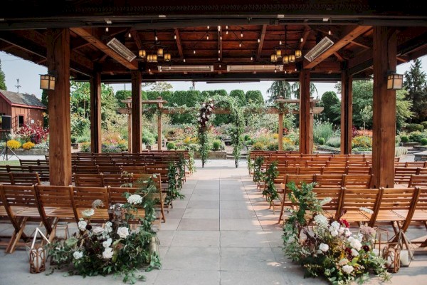 The image shows an outdoor covered wedding setup with wooden chairs and floral decorations, set in a garden under a rustic wooden pergola.