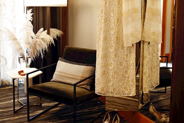 The image shows a room with a lace dress hanging in front of a mirror, a pair of heels on the floor, an armchair, a side table, and a standing lamp.