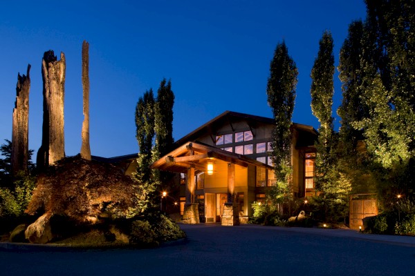 A well-lit building with a rustic design, surrounded by tall trees and lush greenery, set against a clear evening sky.