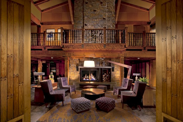 A cozy, rustic living room with a stone fireplace, high ceilings, wooden beams, modern chairs, and warm lighting that creates a welcoming atmosphere.