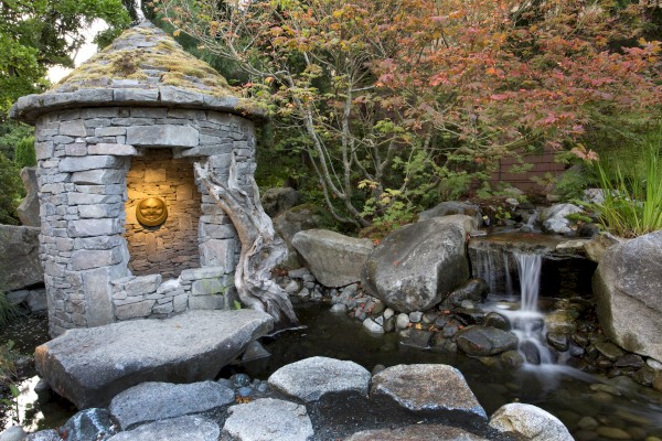 A stone structure with a lit interior beside a small waterfall and pond, surrounded by rocks and trees with autumn foliage, is depicted in the image.