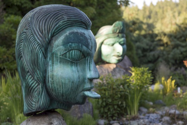 The image shows two large greenish-blue statues of human heads with intricate designs, situated in a lush, outdoor garden setting.