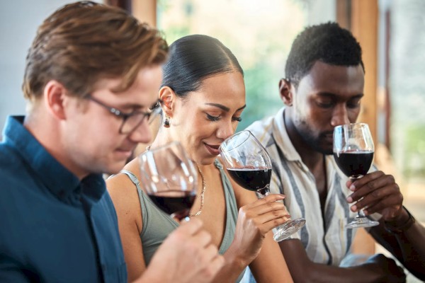 Three people are sitting together, holding glasses of red wine, and appear to be sampling or smelling the wine.