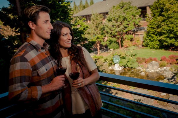 A couple stands on a balcony holding wine glasses, enjoying a landscaped garden view with trees and a building in the background.