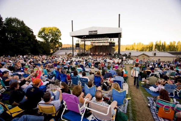 A large crowd is seated on chairs and blankets outdoors, facing a stage with a performance taking place in front of them, under a clear sky.