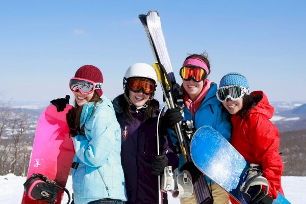 Four people in winter gear standing on a snowy slope, holding snowboards and skis, smiling and ready for snow sports, under a clear sky.