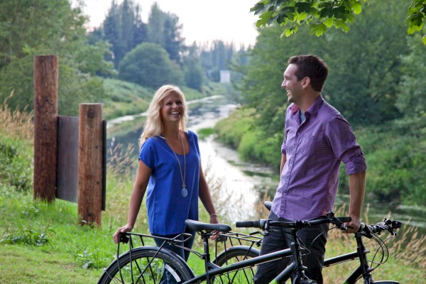 Two people stand with bicycles near a river, smiling and chatting in a green, scenic outdoor area with trees and grass around.