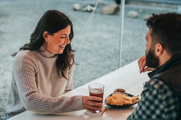 A woman and a man are sitting at a table, the woman holding a glass, and the man with a plate of nachos and dip in front of him.