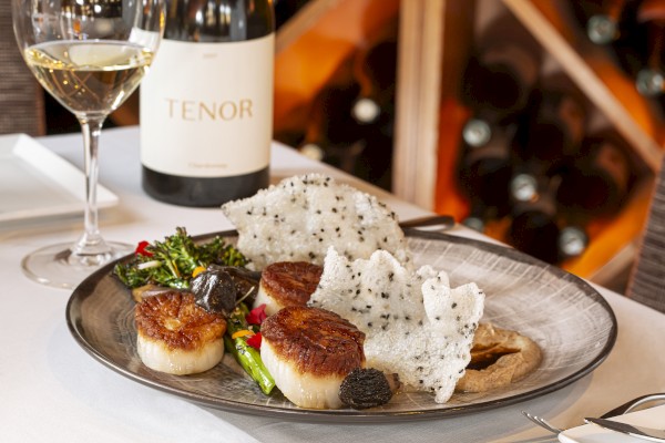 The image shows a plated gourmet meal with seared scallops, vegetables, and crispy white garnishes beside a glass of white wine and a wine bottle.