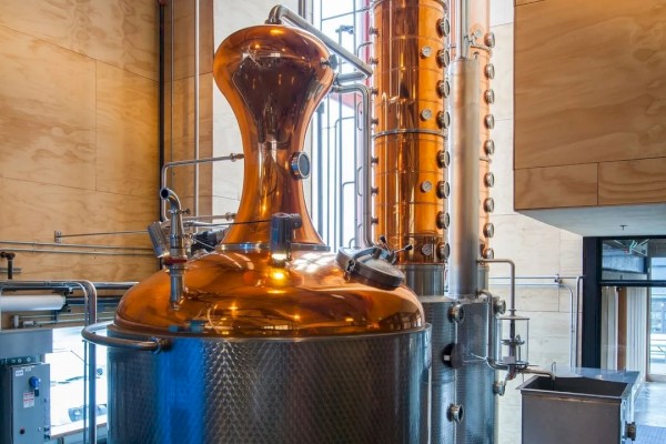 The image shows a distillation apparatus with copper and metal components inside a distillery or brewery setup, featuring pipes and tanks.