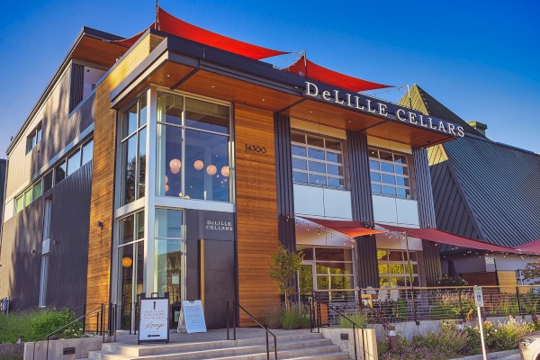 The image shows the exterior of DeLille Cellars, a modern two-story building with wooden accents, large windows, and red shade sails above the entrance.