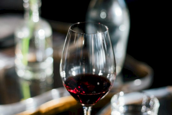 A wine glass filled with red wine is on a reflective table with a vase of flowers, a glass, and a decanter in the background.