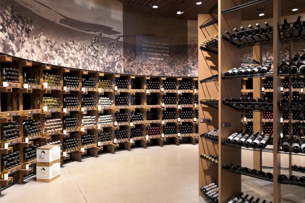 The image shows a well-organized wine store with numerous bottles displayed on shelves arranged in a curved layout and additional shelves in the foreground.