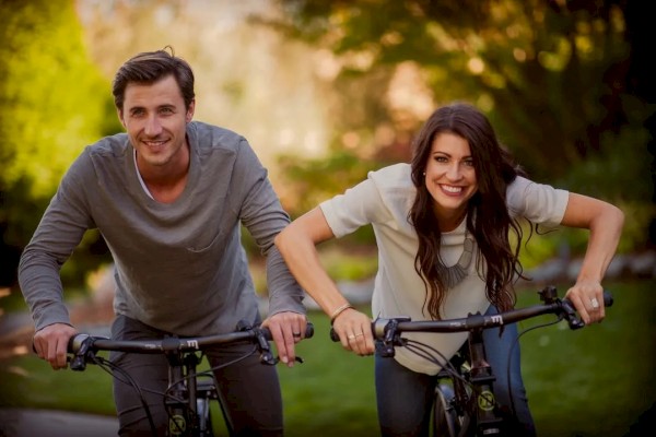 Two people are riding bicycles outdoors, smiling and enjoying a sunny day in a park setting.