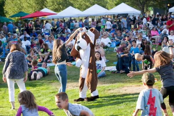 A crowded outdoor event with people dancing and a person in a dog costume. Some are sitting on the grass and there are tents in the background.