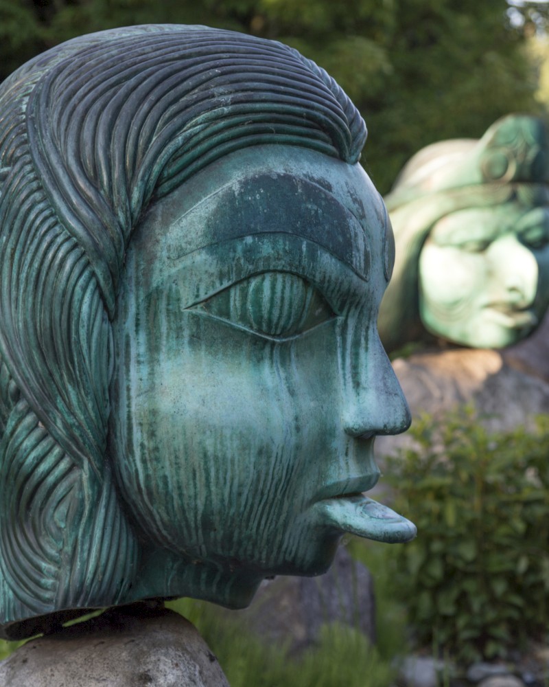 The image shows two large, green-colored sculptures of human-like heads with artistic detailing, positioned outdoors in a lush, green setting.