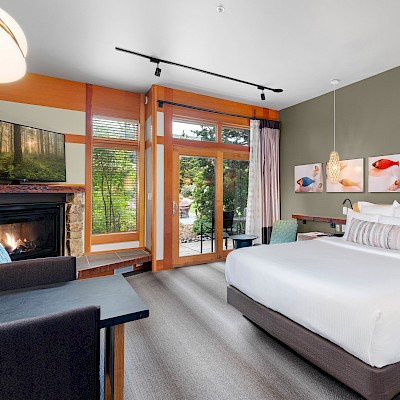 The image shows a modern hotel room with a large bed, artwork on the wall, a fireplace, and a view of greenery through a glass door.