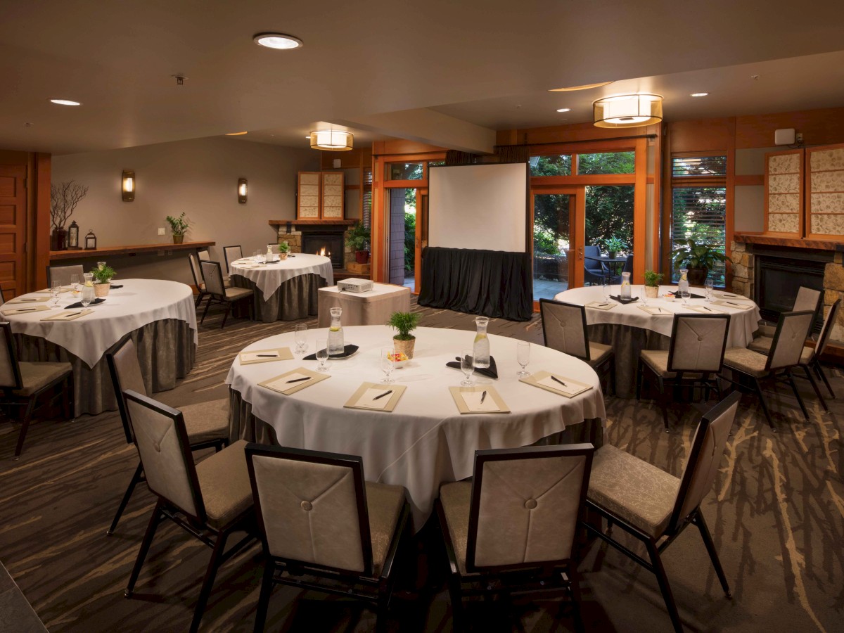 A conference room setup with round tables, chairs, a projector screen, and notepads on tables, lit warmly with natural light from large windows.