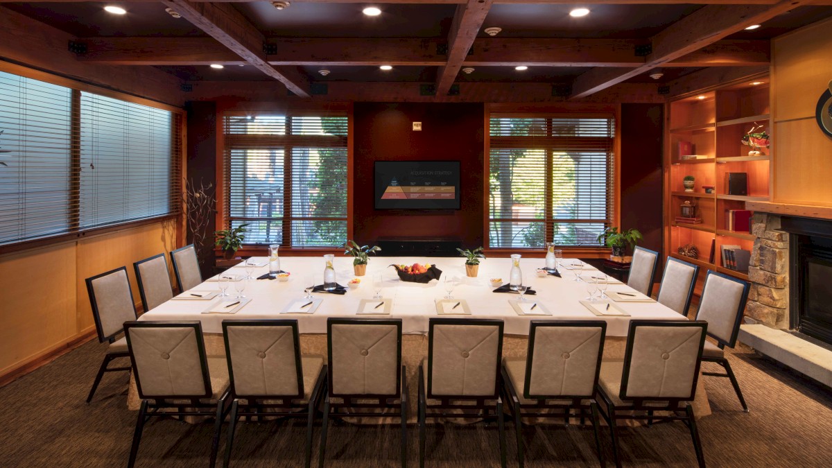 A well-lit conference room with a rectangular table, chairs, a fireplace, bookshelves, and large windows is featured in the image.